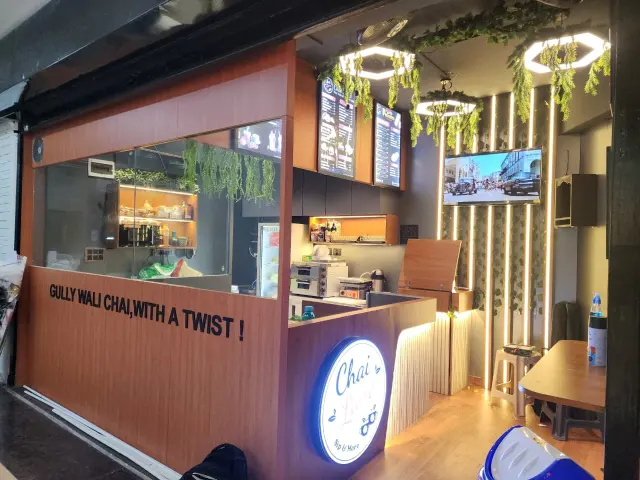 Inside Chai Lane: a wood-panelled counter reading Gully wali chai, with a twist, digital menu boards, hanging greenery and warm strip lights.